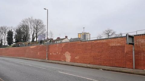 A very long red brick wall next to a road. There is a pavement by the wall and short railings on top. Behind it are houses and a park.