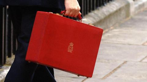 A man's hand is seen holding the Budget red box with the emblem of the late Queen on it. 