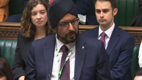 Warinder Juss, wearing a dark suit, stands in the House of Commons, flanked by other MPs sitting behind him
