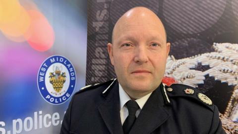 A man in a police uniform stands in front of a poster. He is bald and has blue eyes. The police uniform consists of a black jacket with gold embroidered logos on the shoulders, a white shirt and a black tie. On the poster behind him, a blue and white logo with the words "West Midlands Police" can be seen.