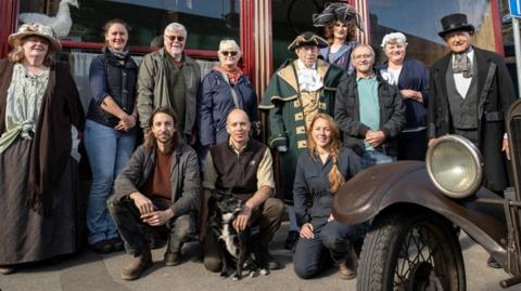 A group of people in Victorian-era outfits stand outside a shop smiling at the camera. There are historic vehicles in the foreground.