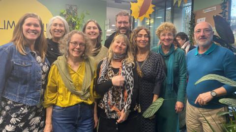 A group of nine people smiling for the camera. They are standing in the climate hub, which has sage green walls and a yellow round logo that reads Climate Hub.