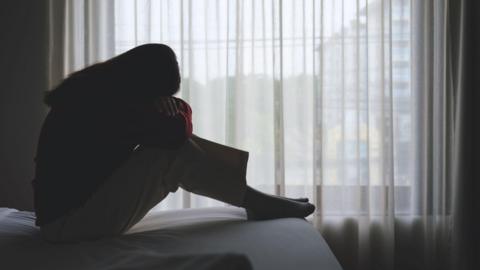 A stock image of a woman sitting and crying on a bed in bedroom. She is in darkness. Some light is coming in from the sheer curtain and some trees and buildings can be seen outside through it.