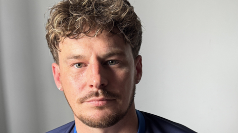 A man with curly brown hair and a thin beard in front of a grey background