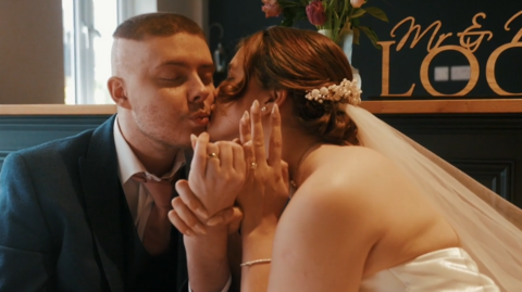 Charlie and Judy kissing on their wedding day. The pair are sat down holding hands. Charlie is in a blue suit and pink tie. Judy is wearing her wedding dress. A wooden sign "Mr & Mrs Lock" is in the background.