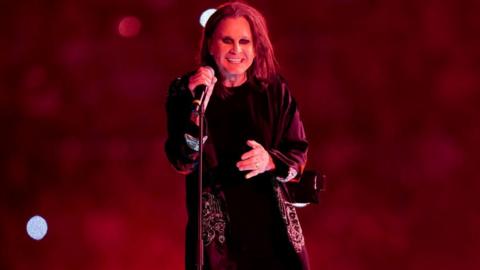 Ozzy Osbourne smiling with a microphone on a stand in his hand. He is wearing a long dark jacket and the background is red with some lights. 