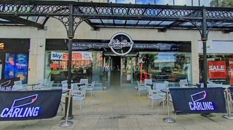 A general view shot of the front entrance of Sky Bar on Kay Gardens in Bury. The bar has a glass front with a black and white logo and branding with seating outside, housed on the outside of a shopping centre. 