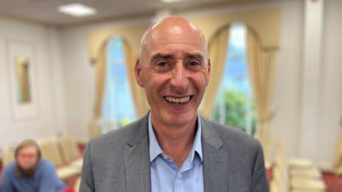 A smiling bald man in a grey suit and blue shirt stands in a room with rows of chairs.