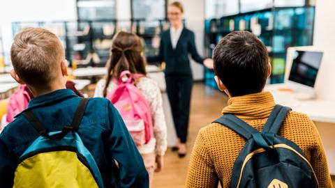 Students being welcomed into a library and computer lab, seen from behind, wearing backpacks.