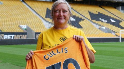 A woman holds an orange football t-shirt with word Cullis and the number 70. She is standing on the pitch at a stadium.