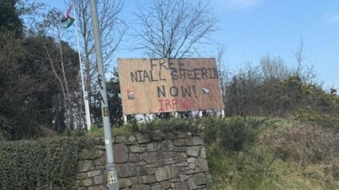 A billboard that says FREE NIALL SHEERIN NOW! IRPWA It is on top of a grassy bank with a stone wall and lampost at the front. 