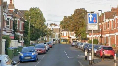 A residential road with cars parked on either side. Large trees can be seen at the far end of the street.