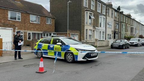 A police officer stands next to a patrol car on a cordoned off street. 