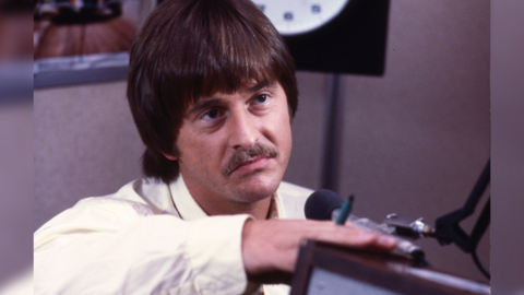 A vintage image of a brown haired man with a 1970s-era bowl haircut and moustache facing a microphone. 