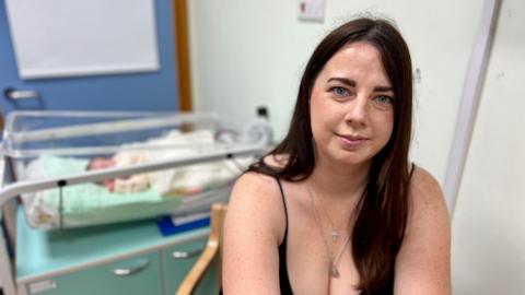 A woman with long brown hair and a dark top is sitting next to her newborn baby, who is asleep in a cot.