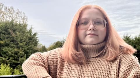 Beatrice Jones sitting on a bench outside. She has pink hair, glasses, and is wearing a pink jumper.