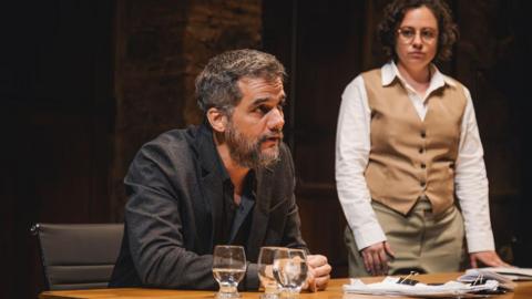 A man sitting at a table with a woman standing beside him. There are glasses containing water on the table and a stack of papers. The man has dark hair and a beard and is wearing a dark blazer over a dark shirt. He is leaning slightly forward with his arms resting on the table. The woman has short, curly hair and is wearing glasses. She is dressed in a white shirt and a tan waistcoat and trousers. 