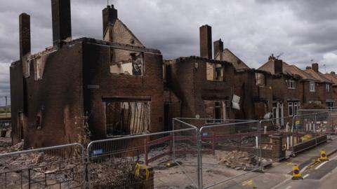 A row of burnt-out semi-detached homes in Wennington in East London in 2022. The brown brickwork is charred, with no roof or windows. There are metal barriers in front.