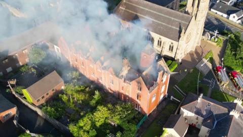 Drone footage from above of a red-brick building with flames inside it. The building is large and long with its roof caved in. It is surrounded by small red-brick buildings and a grey stone church beyond it. Firefighters in hi-viz protective clothing are spraying it with water. 