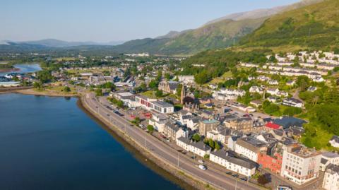 An aerial view of Fort William on a sunny day. The town is on a shore of Loch Linnhe. Rows of houses and other buildings cover a hillside.