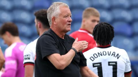 Chris Wilder taps his chest to thank the supporters after the 3-0 win at Mansfield in the EFL Cup. Romelle Donovan, wearing Sheffield United's white away shirt, can be seen behind him with his back turned.