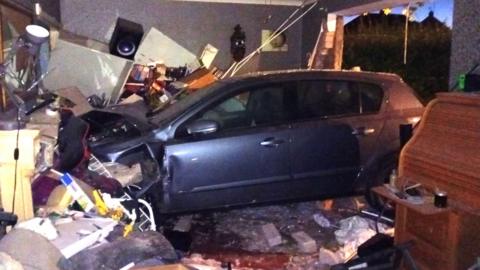 A view inside Alex Richardson's family living room with a car crashed through the front window, with a lot of visible destruction.