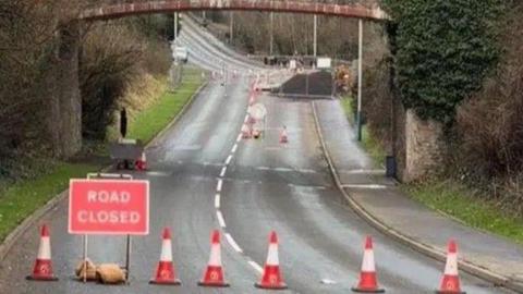 A red Road Closed sign on Kewaigue Hill alongside a line of traffic cones. Works can be seen in the distance and there is a railway bridge over the carriageway.