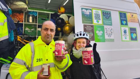 A man and a woman volunteering for Hearty Earth posing for a picture in front of the initiative's van. They are holding cans of red kidney beans in their hands. The man is wearing a yellow high vis outfit. The woman is wearing a helmet.