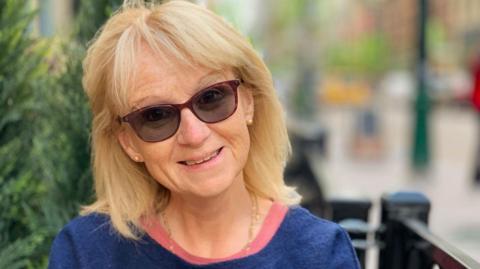 Judith has shoulder length blonde hair, is smiling, and wearing a set of sunglasses. She is wearing a blue jumper on top of a pink shirt.