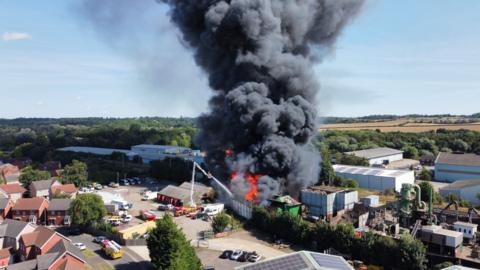 Aerial of the fire at Sackers recycling centre which is surrounded by homes. Flames and a huge plume of black smoke can be seen reaching towards the sky.