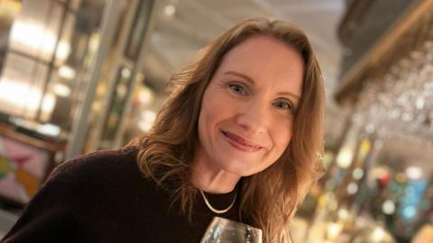 A woman in a burgundy top looking at the camera smiling. She is holding a wine glass and behind her is a out of a focus pub.