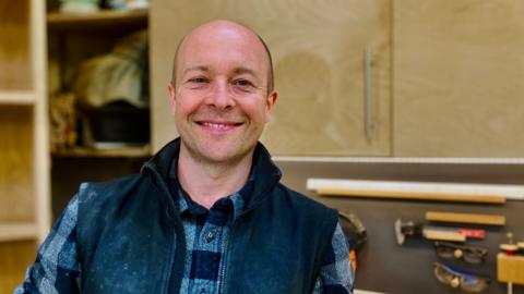 Furniture maker Nick James is in his furniture making workshop in Newcastle.  He is looking at the camera and smiling and wearing his casual clothing. Behind him you can see his wood-working tools on the wall.           