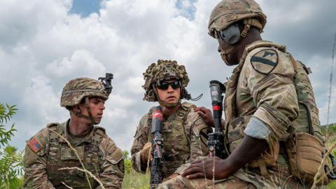 Three US soldiers in a field, wearing helmets and holding weapons and tactical gear