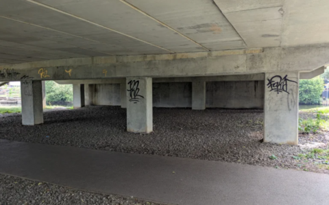 A picture of a bridge underpass. It is grey and concrete with random black graffiti sprayed on concrete pillars.