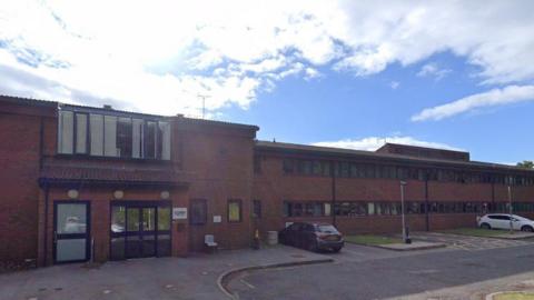 Two storey red brick building with lots of windows to the right of the picture and a set of doors to the left of the building