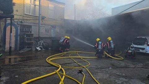 Four firefighters are crouching on a concrete car park in front of a building. They are holding yellow hose jets and are directing water at a building which has smoke coming out of it.