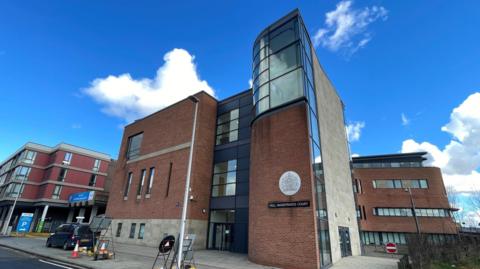 A photo of Hull Magistrates Court building. It has two floors and is a mix of brown brick and clear windows. There is a car parked in front and some roadwork signage and cones.
