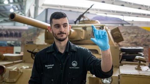 Museum workshops team leader Aaron, who found the round, is holding it between the fingers of his left hand for a picture. Exhibited tanks can be seen behind him.
