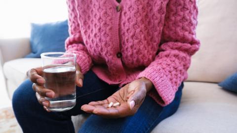 Close-up of a woman wearing a pink jumper sitting on a couch holding medication/supplements in one hand and a glass of water in the other.