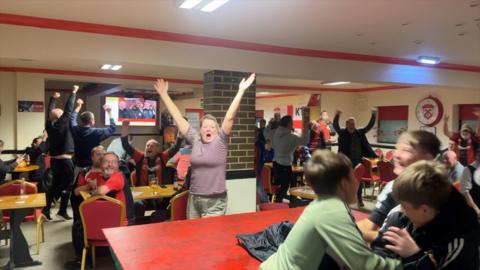 Kettering fans react to their draw agains Northampton. They are inside the clubhouse at the ground, which has tables and chairs. Many fans are jumping with their arms in the air.