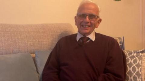 A man with short white hair wearing glasses is smiling and wearing a burgundy jumper with a burgundy tie and white shirt underneath. He is sat on a grey sofa with three grey pillows on it.