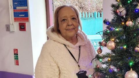 Linda Huskisson wearing a white cable-link jumper standing next to a Christmas tree 