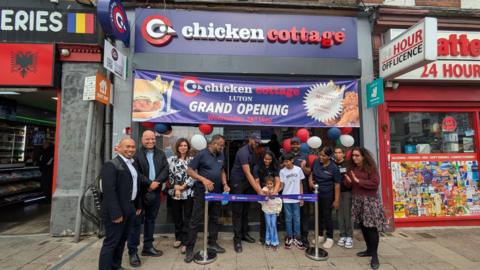 A group of people, some wearing uniforms for a chicken shop, and others smartly dressed, are celebrating outside a new Chicken Cottage chain