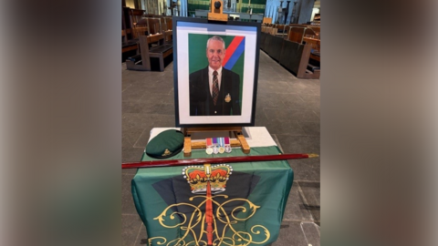 A photo frame standing up with a picture of a man with grey hair wearing a suit in it. In front of it is a small table with military flags, a green beret and medals. It is inside a cathedral