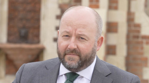 Nick Timothy looks at the camera as he photographed outside. He has facial hair and is wearing a grey suit with a green tie. Behind him is a red-bricked building. 