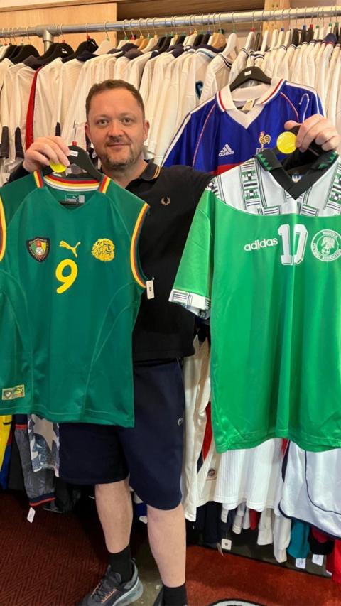A man stands in front of a rack full of England football shirts holding up two football shirts. The shirt on the left is a 2002 Cameroon shirt which is sleevless. It is dark green with a yellow '9' on it. On the right is the 1994 World Cup shirt for Nigeria, it is mostly a light green with a white upper panel with some dark green patterns. 