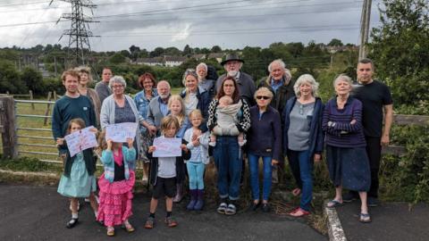 A large group of residents consisting of both adults and children. They are standing in front of the gate that leads into the field where the proposed facility would have been built. The children are holding hand drawn paper signs which say 'save nature'. The sky is grey and cloudy. In the distance you can see a large electricity power line from the neighbouring substation.