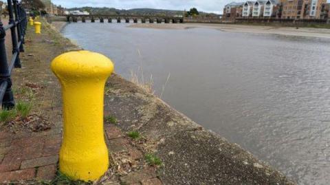 Castle Quay at Barnstaple on a cloudy day. There is a row of yellow mooring bollards along the iron railing of the quayside with one prominent in the foreground. Over the calm water is a row of five-storey housing. Spanning the water in the background is a road bridge.
