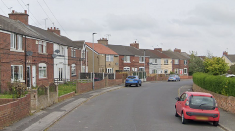 Google streetview image of a residential street with a few cars parked along it. It looks quiet and is a grey day. The houses are terraces and mostly unpainted brick with a few white and beige ones.