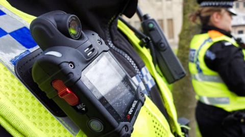The image shows a close-up of a small black box attached to the hi-vis vest of a police officer. A red button on the side has the letters "REC" in white underneath.